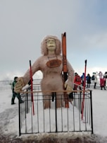A large, furry statue resembling a yeti or snow creature holding skis stands behind a black metal fence. The statue has long hair and is wearing glasses. The background features snow-covered ground and several people in winter clothing, indicating a cold environment. The sky is overcast with a hint of blue.