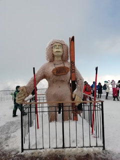 A large, furry statue resembling a yeti or snow creature holding skis stands behind a black metal fence. The statue has long hair and is wearing glasses. The background features snow-covered ground and several people in winter clothing, indicating a cold environment. The sky is overcast with a hint of blue.