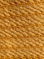 A close-up view of a textured woven surface featuring horizontal rows of tan and light brown fibers intertwined with visible strands. The fibers appear coarse, with occasional hair-like strands protruding, highlighting the natural texture and craftsmanship.