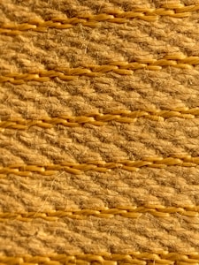 A close-up view of a textured woven surface featuring horizontal rows of tan and light brown fibers intertwined with visible strands. The fibers appear coarse, with occasional hair-like strands protruding, highlighting the natural texture and craftsmanship.