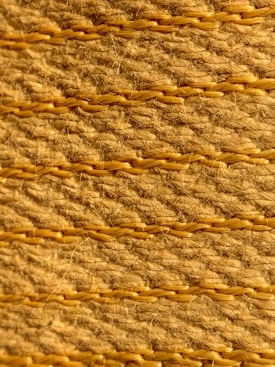 A close-up view of a textured woven surface featuring horizontal rows of tan and light brown fibers intertwined with visible strands. The fibers appear coarse, with occasional hair-like strands protruding, highlighting the natural texture and craftsmanship.