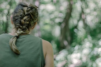 A person with a braided hairstyle, dressed in a green sleeveless top, stands facing a forest background. The setting is lush and green, with soft focus bokeh from trees and foliage.