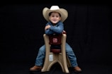 A child wearing a white cowboy hat and denim clothing sits smiling on a beige plastic chair against a black background. The child is wearing brown boots and looks content and confident.