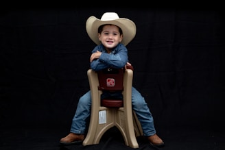 A child wearing a white cowboy hat and denim clothing sits smiling on a beige plastic chair against a black background. The child is wearing brown boots and looks content and confident.