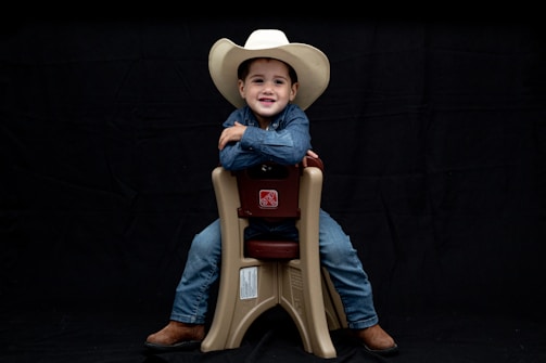 A child wearing a white cowboy hat and denim clothing sits smiling on a beige plastic chair against a black background. The child is wearing brown boots and looks content and confident.
