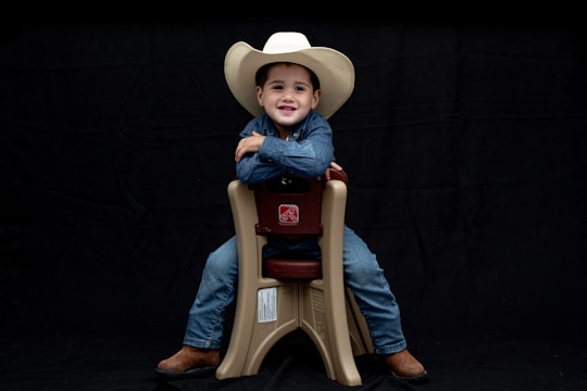 A child wearing a white cowboy hat and denim clothing sits smiling on a beige plastic chair against a black background. The child is wearing brown boots and looks content and confident.