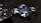 A small, blue quadcopter drone with four propellers is placed on a dark surface. Beside it, there is a black remote control with two joysticks visible. The scene suggests a setup for controlling and flying the drone.