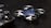 A small, blue quadcopter drone with four propellers is placed on a dark surface. Beside it, there is a black remote control with two joysticks visible. The scene suggests a setup for controlling and flying the drone.