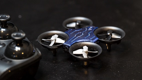 A small, blue quadcopter drone with four propellers is placed on a dark surface. Beside it, there is a black remote control with two joysticks visible. The scene suggests a setup for controlling and flying the drone.