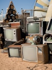 A collection of vintage televisions and other electronic equipment arranged haphazardly on sandy ground. The TVs have wooden casings and bulky screens, reminiscent of mid-20th century designs. Nearby, a retro-style audio speaker system sits alongside various metal and plastic objects. The setup appears to be outdoors, with a clear sky in the background and some modern structures visible.