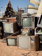 A collection of vintage televisions and other electronic equipment arranged haphazardly on sandy ground. The TVs have wooden casings and bulky screens, reminiscent of mid-20th century designs. Nearby, a retro-style audio speaker system sits alongside various metal and plastic objects. The setup appears to be outdoors, with a clear sky in the background and some modern structures visible.