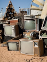 A collection of vintage televisions and other electronic equipment arranged haphazardly on sandy ground. The TVs have wooden casings and bulky screens, reminiscent of mid-20th century designs. Nearby, a retro-style audio speaker system sits alongside various metal and plastic objects. The setup appears to be outdoors, with a clear sky in the background and some modern structures visible.