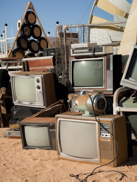A collection of vintage televisions and other electronic equipment arranged haphazardly on sandy ground. The TVs have wooden casings and bulky screens, reminiscent of mid-20th century designs. Nearby, a retro-style audio speaker system sits alongside various metal and plastic objects. The setup appears to be outdoors, with a clear sky in the background and some modern structures visible.