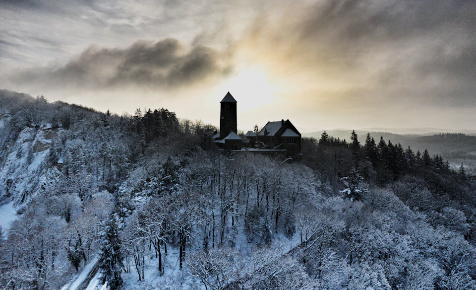 The image depicts a serene winter scene atop a hill, with a tall tower standing out against the backdrop of a cloudy sky and snow-covered landscape. The tower is part of a larger structure that includes a house nestled among the trees. The photograph appears to have been taken from an aerial perspective, providing a comprehensive view of the surrounding area.