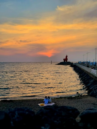 A couple sits on a sandy beach sharing a serene moment as the sun sets, casting warm hues across the sky and reflecting on the calm ocean waters. A red lighthouse stands prominently on a rocky pier, while streetlamps line the walkway.