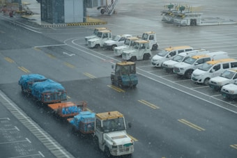 Several airport vehicles, including baggage carts covered with blue tarps, are lined up on the wet tarmac. Snow is falling, and a fleet of white service vehicles with yellow details is parked nearby.