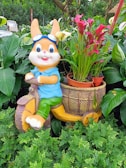 A playful rabbit statue wearing blue goggles and a blue shirt sits on a yellow scooter with a basket carrying pink calla lilies. The background is lush with various green plants, creating a vibrant garden setting.