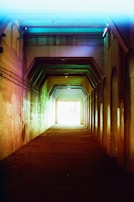 A dimly lit tunnel with geometric patterns created by angular supports along the ceiling. The walls are colored with a gradient of muted colors, including shades of reds, greens, and blues. The tunnel leads towards a bright light at the far end, creating a contrast between the dark interior and the bright exterior visible in the distance.