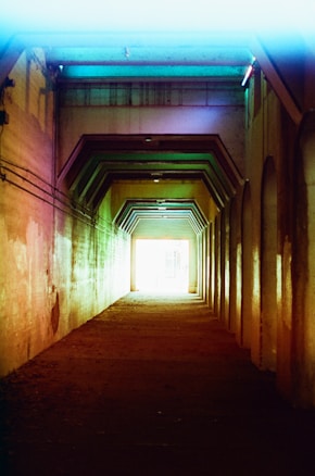 A dimly lit tunnel with geometric patterns created by angular supports along the ceiling. The walls are colored with a gradient of muted colors, including shades of reds, greens, and blues. The tunnel leads towards a bright light at the far end, creating a contrast between the dark interior and the bright exterior visible in the distance.