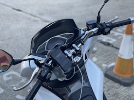 The image shows the handlebars and dashboard of a white motorcycle parked on a paved surface. The throttle and brake controls are visible, as well as a mounted phone holder. Part of a traffic cone can be seen in the background.