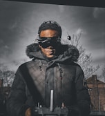 A person wearing a dark parka with a fur-lined hood and a cap is holding a remote control device. The individual is also wearing futuristic goggles with antennas. The background features a cloudy, overcast sky and bare trees, suggesting a cold environment.