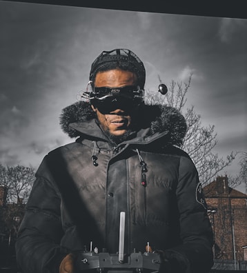 A person wearing a dark parka with a fur-lined hood and a cap is holding a remote control device. The individual is also wearing futuristic goggles with antennas. The background features a cloudy, overcast sky and bare trees, suggesting a cold environment.