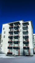 A modern multi-story apartment building with large glass balconies and red accents stands against a deep blue sky. The building's facade features a mix of white and grey panels, and is surrounded by a layer of snow on the ground.