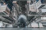 A large aircraft is positioned inside a vast, industrial hangar. The structure is composed of metallic scaffolding and platforms, with maintenance equipment surrounding the aircraft. The perspective is from the ground, highlighting the immense size of both the plane and the hangar.