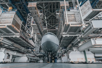A large aircraft is positioned inside a vast, industrial hangar. The structure is composed of metallic scaffolding and platforms, with maintenance equipment surrounding the aircraft. The perspective is from the ground, highlighting the immense size of both the plane and the hangar.
