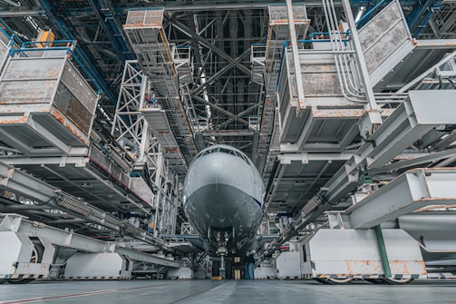 A large aircraft is positioned inside a vast, industrial hangar. The structure is composed of metallic scaffolding and platforms, with maintenance equipment surrounding the aircraft. The perspective is from the ground, highlighting the immense size of both the plane and the hangar.