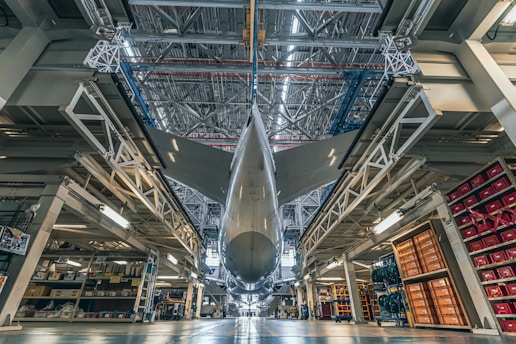 A large aircraft is stationed inside a spacious hangar with its massive structure dominating the scene. The view is from underneath, highlighting the intricate engineering and the expansive wingspan. The hangar is filled with various equipment and storage areas, with shelves and tools surrounding the airplane.