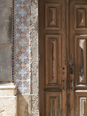 An old wooden door with peeling paint stands next to a wall decorated with intricate blue and orange patterned tiles. The tiles have a traditional design, adding a cultural element to the scene. The door appears weathered, indicating age and character.