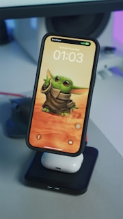 A smartphone displaying an animated character with large ears and green skin, wearing a brown robe, set against a sandy, desert-like background. The phone is placed on a charging stand, with earbuds visible underneath it. The time on the phone reads 01:03, with a date written in Turkish.