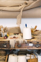 A cluttered workbench with various weaving materials and tools. Rolls of white thread, large rolls of burlap fabric, and completed woven items are prominently displayed. The surface is littered with thread scraps, a small wooden loom, and assorted weaving accessories. Baskets and containers holding more supplies are stored underneath and on the bench.