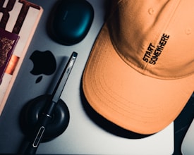 A close-up view of several items on a flat surface, including an orange baseball cap with the words 'Start Somewhere' embroidered on it, a stylus pen on a small black stand, a partially visible electronic device, an Apple logo, and another tech accessory.