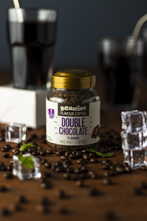 A jar labeled 'Beanies Flavor Coffee Double Chocolate' is surrounded by coffee beans and pieces of mint leaves scattered on a wooden surface. Two glasses filled with a dark liquid, possibly coffee, are seen in the background, along with a few ice cubes placed around the jar.