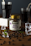 A jar labeled 'Beanies Flavor Coffee Double Chocolate' is surrounded by coffee beans and pieces of mint leaves scattered on a wooden surface. Two glasses filled with a dark liquid, possibly coffee, are seen in the background, along with a few ice cubes placed around the jar.