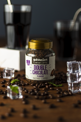 A jar labeled 'Beanies Flavor Coffee Double Chocolate' is surrounded by coffee beans and pieces of mint leaves scattered on a wooden surface. Two glasses filled with a dark liquid, possibly coffee, are seen in the background, along with a few ice cubes placed around the jar.