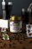A jar labeled 'Beanies Flavor Coffee Double Chocolate' is surrounded by coffee beans and pieces of mint leaves scattered on a wooden surface. Two glasses filled with a dark liquid, possibly coffee, are seen in the background, along with a few ice cubes placed around the jar.