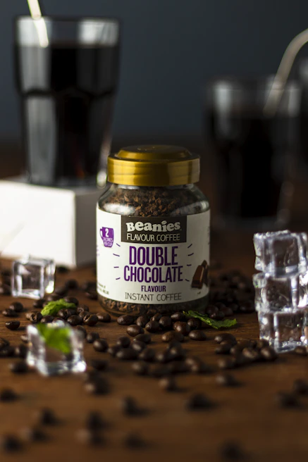 A jar labeled 'Beanies Flavor Coffee Double Chocolate' is surrounded by coffee beans and pieces of mint leaves scattered on a wooden surface. Two glasses filled with a dark liquid, possibly coffee, are seen in the background, along with a few ice cubes placed around the jar.