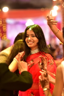A woman is smiling amidst a lively atmosphere, adorned in a traditional red saree with floral embroidery and a choker necklace. Her hair is loose, and she is surrounded by people whose hands are visible, suggesting a celebratory event. The background is illuminated with warm lights and vibrant colors, adding to the festive ambiance.