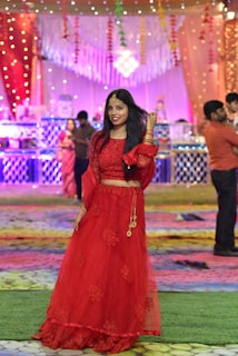 A woman wearing a traditional red outfit stands on a colorful patterned mat. She is smiling and holding her hair. The background is decorated with string lights and drapery, suggesting a festive or celebratory setting. Several people can be seen in the background, along with tables and other decorations.