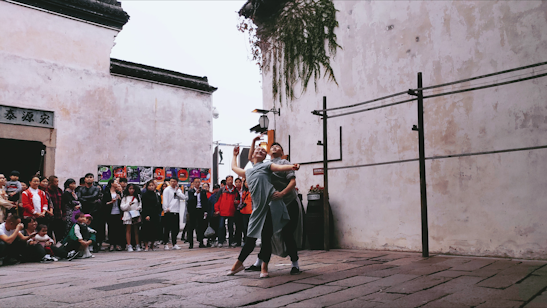 Two performers engage in a contemporary dance routine on a cobblestone street, surrounded by an audience. The scene takes place in an urban setting with traditional architectural elements, including a faded wall and overhead greenery.
