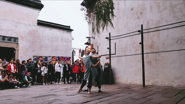 Two performers engage in a contemporary dance routine on a cobblestone street, surrounded by an audience. The scene takes place in an urban setting with traditional architectural elements, including a faded wall and overhead greenery.