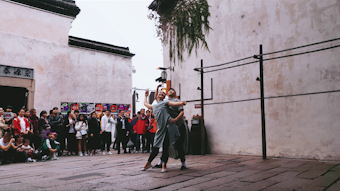 Two performers engage in a contemporary dance routine on a cobblestone street, surrounded by an audience. The scene takes place in an urban setting with traditional architectural elements, including a faded wall and overhead greenery.