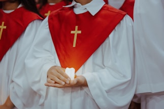 A person dressed in a white robe with a red stole featuring a gold cross is holding a lit candle. The focus is on the hands and the candle, with other similarly dressed individuals in the background.