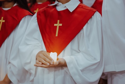 A person dressed in a white robe with a red stole featuring a gold cross is holding a lit candle. The focus is on the hands and the candle, with other similarly dressed individuals in the background.