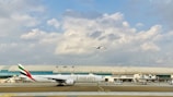 An airport scene with a large Emirates airplane on the runway and another aircraft in the sky above. The background features a modern airport terminal with large windows and a partly cloudy sky.