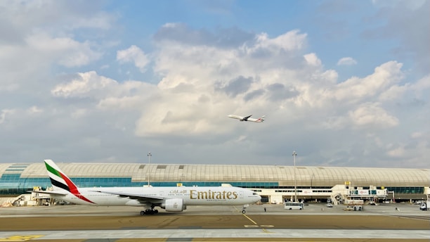 An airport scene with a large Emirates airplane on the runway and another aircraft in the sky above. The background features a modern airport terminal with large windows and a partly cloudy sky.