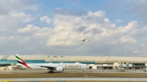 An airport scene with a large Emirates airplane on the runway and another aircraft in the sky above. The background features a modern airport terminal with large windows and a partly cloudy sky.